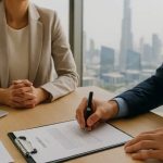 Two business professionals sit at a desk with documents as one signs an agreement in a modern office setting.