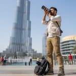 Photographer capturing cityscape near Burj Khalifa in Dubai, standing with camera and backpack as people walk and fountains flow in background